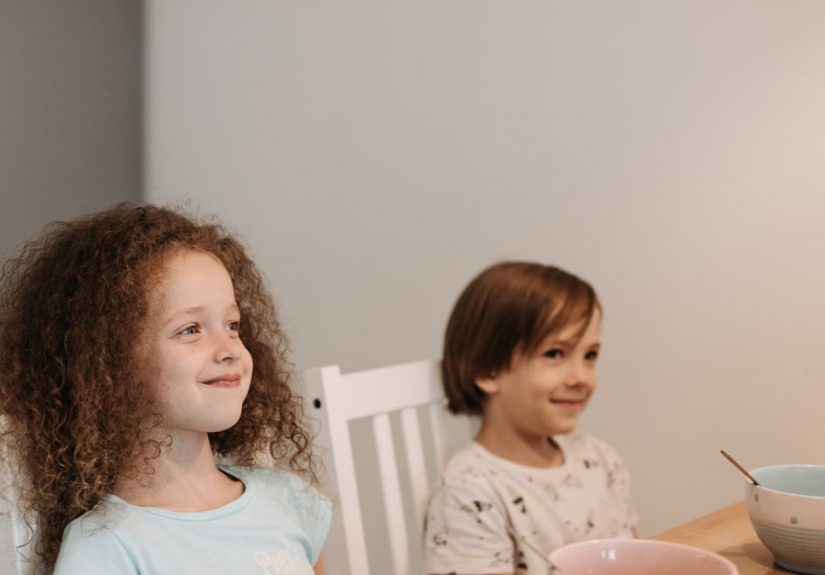 Two children sitting at a table smiling, enjoying their breakfast time indoors.
