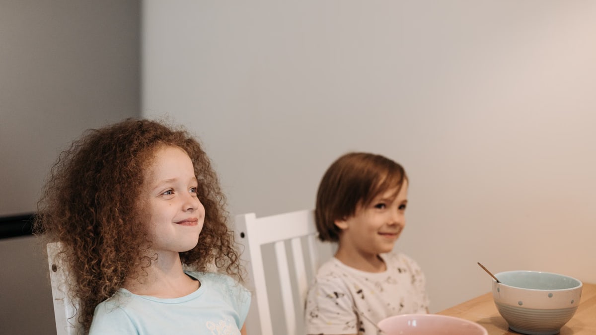Two children sitting at a table smiling, enjoying their breakfast time indoors.
